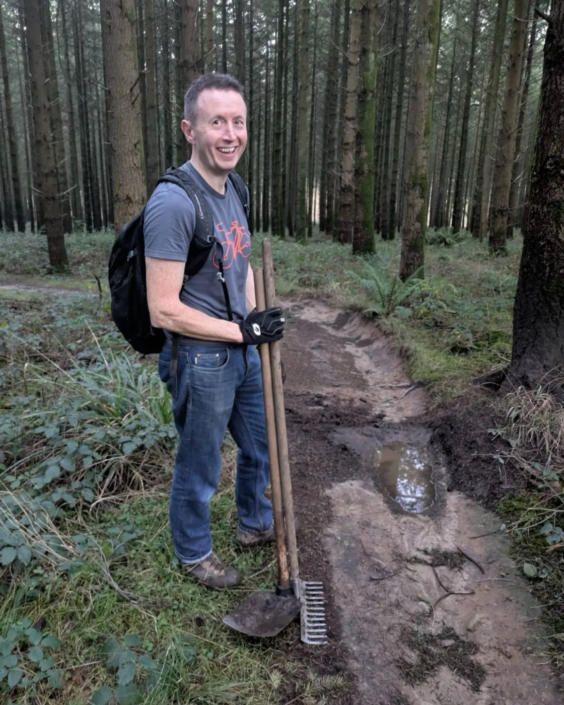 Sam Burrough on a woodland bike trail in the Forest of Dean, holding a shovel and rake